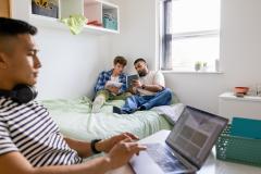 Un étudiant travaille sur son bureau pendant que deux autres révisent assis sur le lit d'une chambre d'étudiant