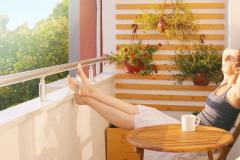 Une jeune femme assise sur son balcon avec les pieds relevés contre la rambarde profite du soleil, les yeux fermés