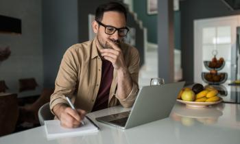 homme, installé dans sa cuisine, regarde son ordinateur un crayon à la main
