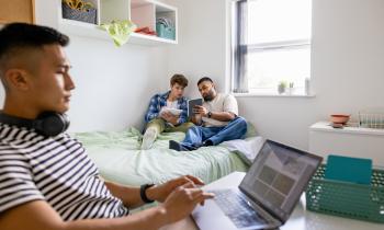 Un étudiant travaille sur son bureau pendant que deux autres révisent assis sur le lit d'une chambre d'étudiant