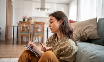 Jeune femme assise à terre contre son canapé dans le salon, et prenant des notes. 