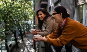 Deux femmes brunes rigolent, une tasse de café à la main, sur un balcon. 
