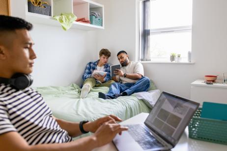 Un étudiant travaille sur son bureau pendant que deux autres révisent assis sur le lit d'une chambre d'étudiant
