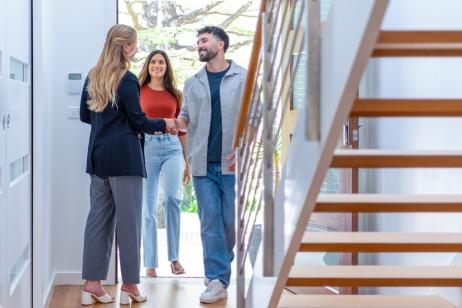 Un jeune couple est accueilli sur le pas de la porte par une femme aux cheveux longs blonds pour visiter une maison