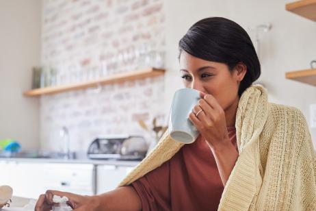 Working from home, a woman sick with covid virus or flu and doing business email with laptop on the kitchen table. Remote office work, online employee has tea and blanket to stay warm in poor health
