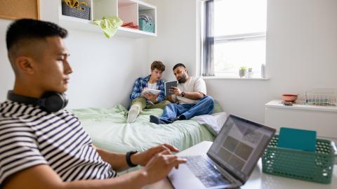 Un étudiant travaille sur son bureau pendant que deux autres révisent assis sur le lit d'une chambre d'étudiant