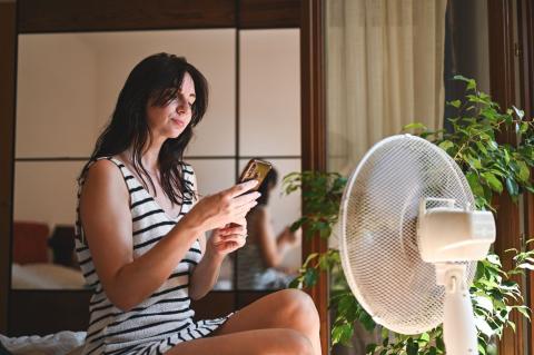 Jeune femme assise devant sa fenêtre, près d'un ventilateur et d'une plante verte, regarde son téléphone