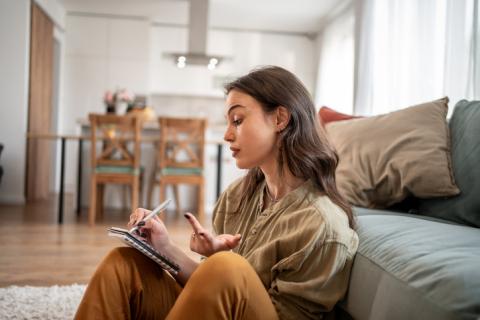 Jeune femme assise à terre contre son canapé dans le salon, et prenant des notes.