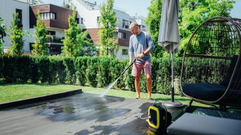 Un homme en bermuda et t-shirt nettoie sa terrasse en bois à l'aide d'un jet d'eau à haute pression