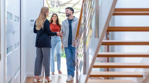 Un jeune couple est accueilli sur le pas de la porte par une femme aux cheveux longs blonds pour visiter une maison