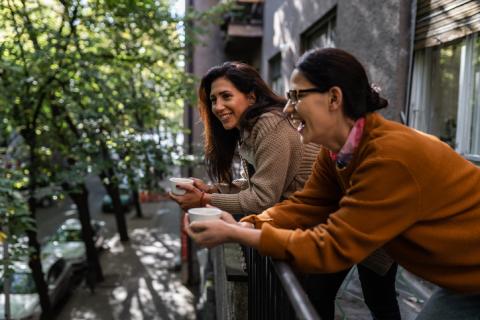 Deux femmes brunes rigolent, une tasse de café à la main, sur un balcon. 