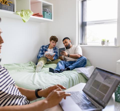 Un étudiant travaille sur son bureau pendant que deux autres révisent assis sur le lit d'une chambre d'étudiant