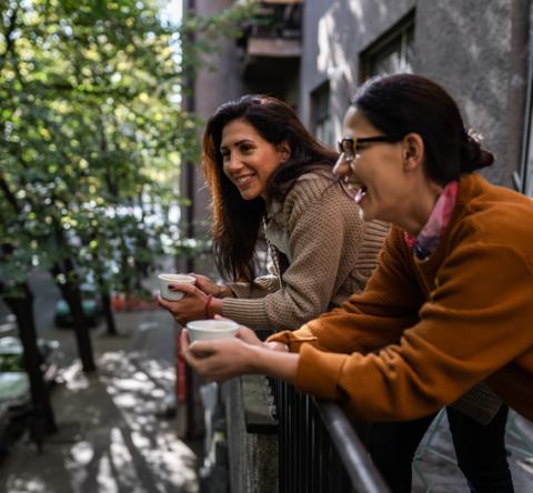 Deux femmes brunes rigolent, une tasse de café à la main, sur un balcon. 
