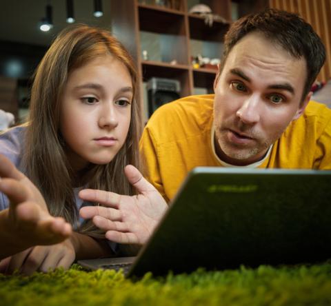 Un père et sa fille adolescente regardent un écran d'ordinateur, allongés à terre. 